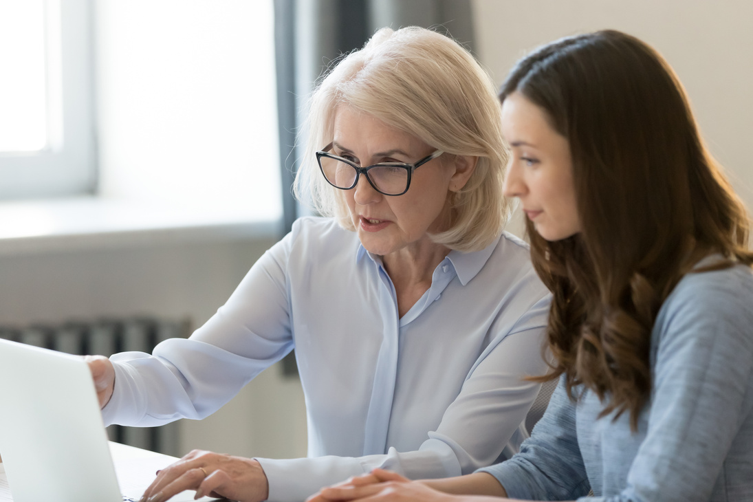 Serious old mentor teaching intern or student pointing at laptop